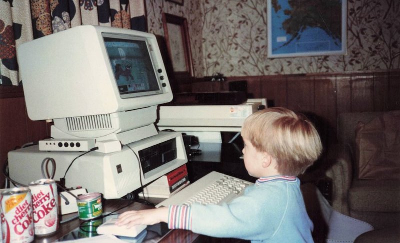 A young child sitting at a large beige IBM PC computer that towers above them, using an early computer mouse while looking at a CRT monitor displaying what appears to be an educational program.