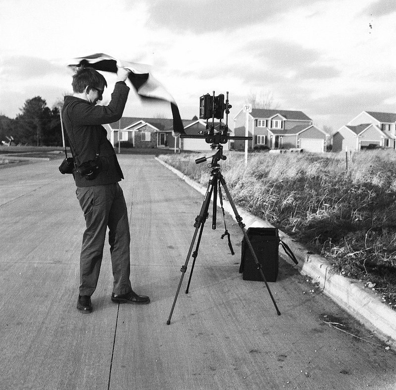 A person crouched behind a large format camera on a tripod in the middle of a residential street, with houses and trees visible in the background.
