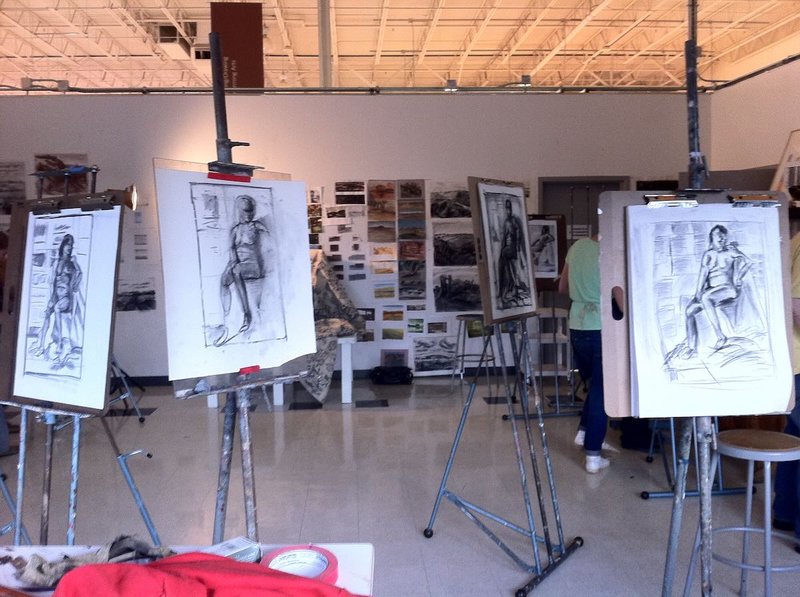 A large open studio space with exposed ceiling beams and industrial lighting, containing multiple easels with charcoal figure drawings, art supplies scattered on tables, and a person in green working at an easel in the background.